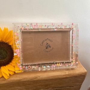 Può includere: Una cornice rettangolare trasparente con coriandoli colorati e le parole "LOVE", "LAUGH" e "HOME" incorporate nella cornice. La cornice ha un retro marrone e poggia su una superficie di legno accanto a un girasole.