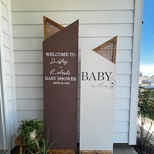 Può includere: Due cartelli in legno con sfondo marrone e bianco. Il cartello a sinistra dice "Benvenuti a Destiny + Richards Baby Shower June 29, 2024". Il cartello a destra dice "Baby in Bloom" con un design floreale.