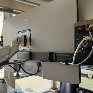 May include: A rear view of a computer monitor mounted on an arm with a small wooden speaker mounted to the right of the monitor. The speaker has a black face with a volume knob and several ports. The speaker is connected to the monitor with wires.
