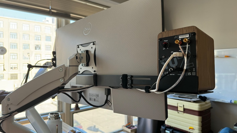 May include: A rear view of a computer monitor mounted on an arm with a small wooden speaker mounted to the right of the monitor. The speaker has a black face with a volume knob and several ports. The speaker is connected to the monitor with wires.