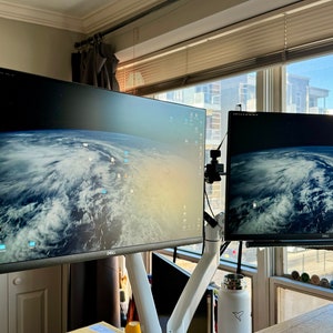 May include: Two computer monitors with a desktop background of a cloudy Earth. The monitors are mounted on a white arm. A pair of wooden speakers are mounted on either side of the monitors.
