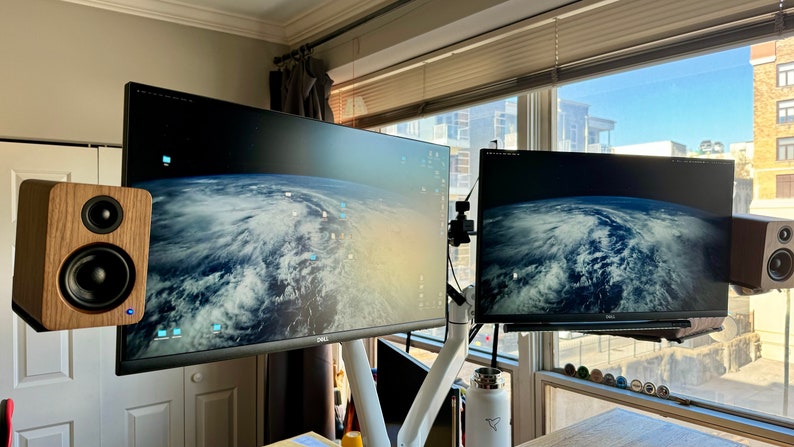 May include: Two computer monitors with a desktop background of a cloudy Earth. The monitors are mounted on a white arm. A pair of wooden speakers are mounted on either side of the monitors.