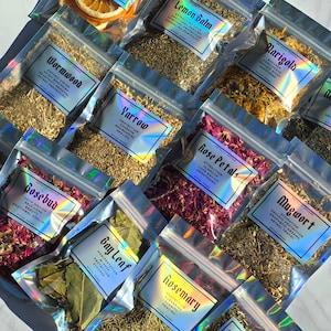 May include: An assortment of dried herbs and botanicals in clear, resealable bags. Each bag features a label with a holographic effect, detailing the herb's name and properties. Herbs include Lemon Balm, Yarrow, and Bay Leaf. The image has a bright, natural light.
