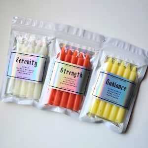 May include: Three clear resealable pouches containing ritual candles. The left pouch holds white candles labeled Serenity, the center holds orange candles labeled Strength, and the right holds yellow candles labeled Radiance. Each label features holographic text.