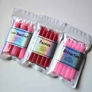 May include: Three sealed plastic bags containing ritual candles. The bags are labeled "Love Manifest", "Passion", and "Attraction". The candles are pink and red. The labels have holographic accents and include keywords like "Romance", "Desire", and "Love".