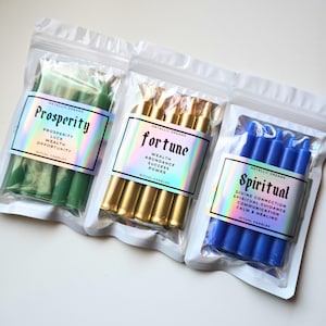 May include: Three sealed plastic bags containing ritual candles. One bag holds green candles labeled "Prosperity", another holds gold candles labeled "Fortune", and the third contains blue candles labeled "Spiritual". Each bag has a holographic label.