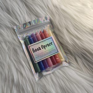 May include: A set of seven thin, multi-colored taper candles in a clear, iridescent plastic pouch. The label reads Metallic Dreams Road Opener, with text explaining the use to remove obstacles and open roads ahead for luck and opportunity.