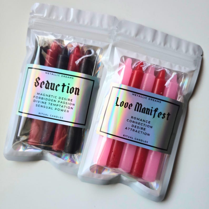 May include: Two clear plastic pouches containing ritual candles. One pouch has red and black candles with a label that reads "Seduction." The other pouch has pink and red candles with a label that reads "Love Manifest." Both labels have the words "Metallic Dreams" and "Ritual Candles."