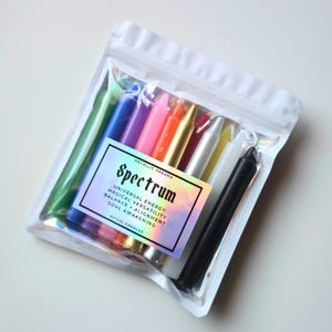 May include: A set of ten multi-colored taper candles in a clear plastic pouch. The label reads: METALLIC DREAMS SPECTRUM UNIVERSAL ENERGY MAGICAL VERSATILITY BALANCE + ALIGNMENT SOUL AWAKENING RITUAL CANDLES. The candles are green, blue, purple, pink, orange, gold, red, silver, yellow, white, and black.