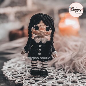 May include: A small crocheted doll with black yarn hair styled in two long braids. The doll wears a black dress with a white collar, white cuffs, and three white buttons, paired with black and white striped leggings and black shoes.