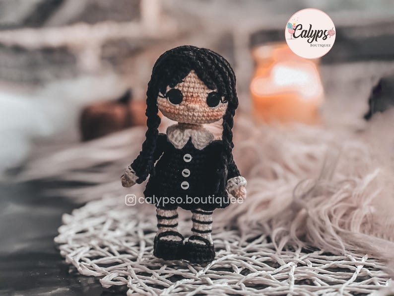 May include: A small crocheted doll with black yarn hair styled in two long braids. The doll wears a black dress with a white collar, white cuffs, and three white buttons, paired with black and white striped leggings and black shoes.