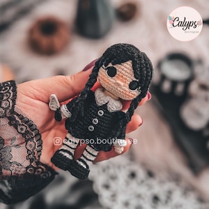 May include: A small crochet doll featuring black hair styled in two long braids, a black dress with a white collar and three small buttons, and black and white striped legs. The doll has a light skin tone and large black eyes. The text calypso.boutique is visible.