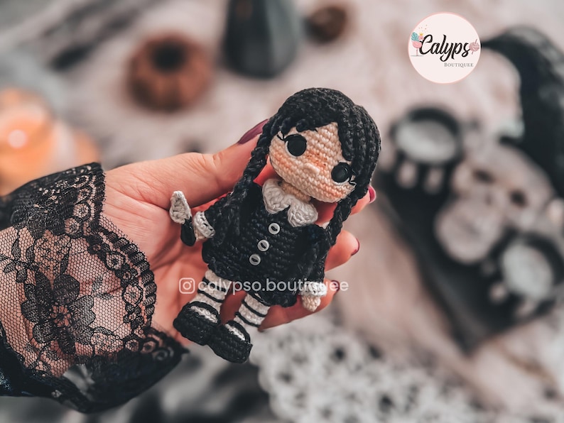 May include: A small crochet doll featuring black hair styled in two long braids, a black dress with a white collar and three small buttons, and black and white striped legs. The doll has a light skin tone and large black eyes. The text calypso.boutique is visible.