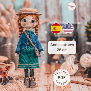 Op de afbeelding: Een handgemaakte gehaakte pop, ongeveer 26 cm hoog, met rood gevlochten haar, gekleed in een blauw jasje en groene rok, en een strohoed. De pop heeft een kleine tas en staat naast een kleine gehaakte hond. De tekst "Anne pattern 26 cm" is zichtbaar.