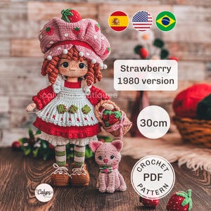 Puede incluir: Una muñeca de ganchillo de 30 cm con pelo de lana roja, que lleva un sombrero con estampado de fresas, un vestido rojo y un delantal blanco. La muñeca sostiene una cesta de fresas y está junto a un pequeño gato de ganchillo rosa. Texto: Strawberry 1980 version.