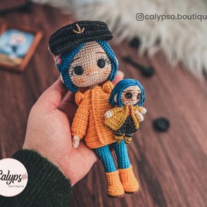 May include: Two crocheted dolls, one larger than the other, are wearing yellow raincoats and blue trousers. The larger doll has blue hair and a black hat with an anchor on it. The smaller doll has blue hair and is holding a yellow raincoat. The dolls are on a wooden surface.