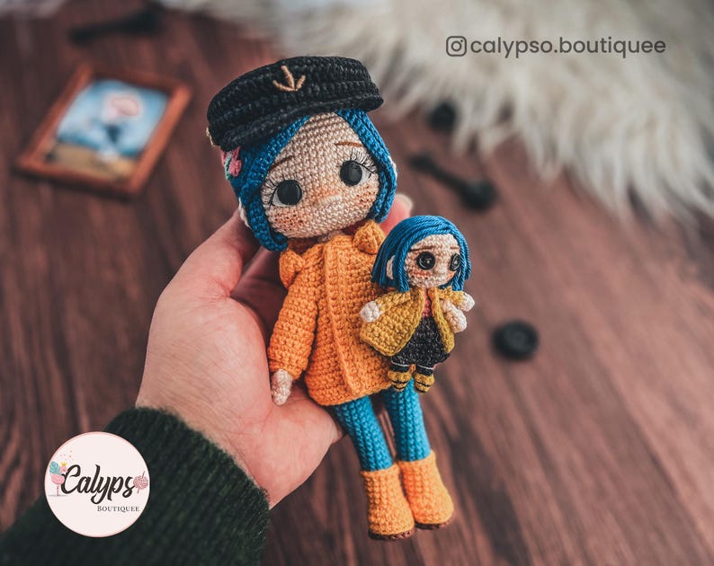 May include: Two crocheted dolls, one larger than the other, are wearing yellow raincoats and blue trousers. The larger doll has blue hair and a black hat with an anchor on it. The smaller doll has blue hair and is holding a yellow raincoat. The dolls are on a wooden surface.