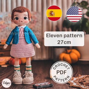 Puede incluir: Muñeca de ganchillo de un personaje con vestido rosa y chaqueta azul, de 27 cm de altura. La muñeca lleva zapatillas y calcetines a rayas. Se muestra el texto "Eleven pattern". También es visible un logotipo de "Crochet PDF Pattern".