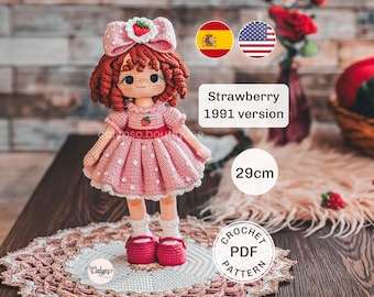 May include: A 29cm tall crocheted doll with reddish-brown curly hair, wearing a pink dress with white polka dots and a matching bow featuring a strawberry emblem. The doll has dark eyes, a small strawberry on the bodice, white socks, and pink shoes.