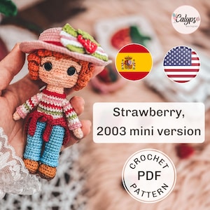 Puede incluir: Muñeca de ganchillo con pelo naranja, que lleva un sombrero rosa con un adorno de fresa, una camisa a rayas, pantalones azules y un cinturón rojo. La muñeca se sostiene en una mano. El texto de la imagen dice "Strawberry, 2003 mini version".