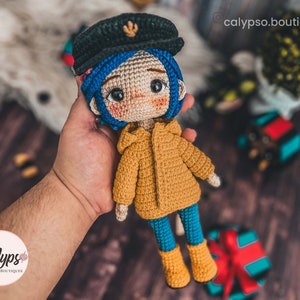 May include: A crocheted doll with blue hair, wearing a yellow jacket, blue trousers, and black boots. The doll is holding a small, crocheted object in its hand.