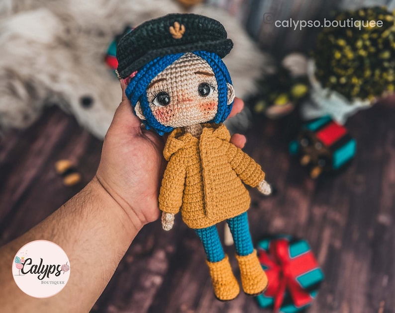 May include: A crocheted doll with blue hair, wearing a yellow jacket, blue trousers, and black boots. The doll is holding a small, crocheted object in its hand.