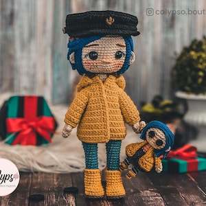 May include: Two crocheted dolls, one larger than the other, both wearing yellow raincoats and blue trousers. The larger doll has a black hat and blue hair. The smaller doll has blue hair. Both dolls have large eyes and are holding hands.