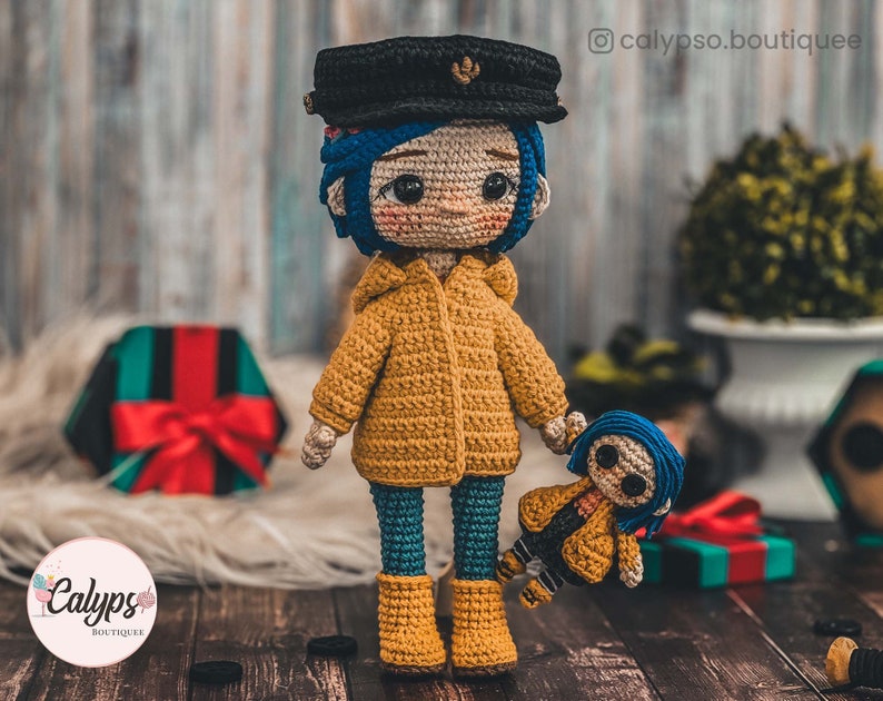 May include: Two crocheted dolls, one larger than the other, both wearing yellow raincoats and blue trousers. The larger doll has a black hat and blue hair. The smaller doll has blue hair. Both dolls have large eyes and are holding hands.