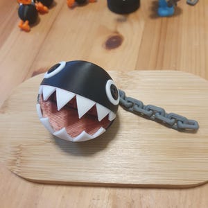 May include: A black and white Chain Chomp character with a gray chain, resting on a wooden surface. The character has a round body, white teeth, and a red interior. Other small figurines are in the background.