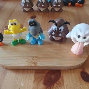 May include: A selection of colourful, miniature figurines of characters from a popular video game franchise. The figures include a yellow bird-like character, a grey turtle, a brown mushroom, a white ghost, and a black bomb. They are displayed on a wooden tray.