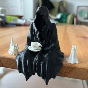Puede incluir: Una figura negra de la Parca con una taza y un platillo blancos, sentada sobre una superficie de madera. También hay dos figuras de gato fantasma blancas y una figura de vestido fantasma blanco. La escena tiene un tema caprichoso y espeluznante.