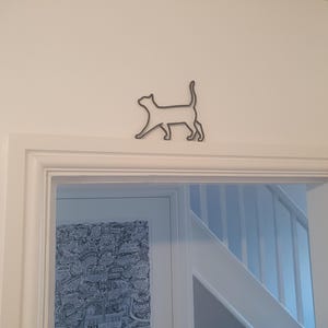 Peut inclure: Décoration murale en forme de silhouette de chat en métal foncé, montée au-dessus d'un cadre de porte blanc. Le chat est représenté en train de marcher. Le fond est un mur blanc et un aperçu d'une pièce.