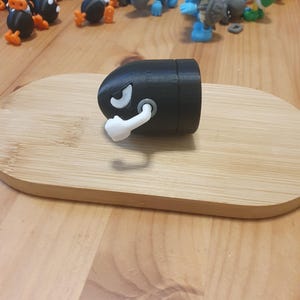 Puede incluir: Una figura negra de Bala de Cañón con una cara blanca y un escape blanco, apoyada sobre un óvalo de madera. Otras figuras coloridas de personajes de videojuegos están en el fondo.