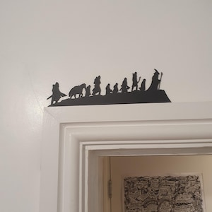 May include: A black metal silhouette of characters from a fantasy story, mounted above a doorway. The figures include a wizard with a staff, and other characters. The artwork is mounted on a white wall.