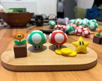 May include: A collection of colorful Super Mario Bros. themed figurines, including green and red mushrooms, a yellow star, a flower, and gold coins, all arranged on a wooden tray. Other figurines are in the background.