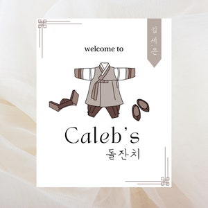 Puede incluir: Una tarjeta blanca con una ilustración marrón y blanca de un atuendo tradicional coreano, zapatos y un sombrero. El texto "welcome to Caleb's 돌잔치" está impreso en la tarjeta.
