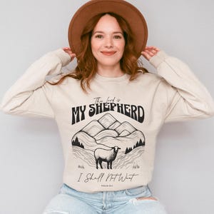 Peut inclure: Un sweat-shirt beige avec un imprimé graphique noir représentant un paysage de montagne et un mouton. Le texte "The Lord is My Shepherd" et "I Shall Not Want" est imprimé sur le sweat-shirt. Le texte "Psalm 23:1" est imprimé sous le graphique.