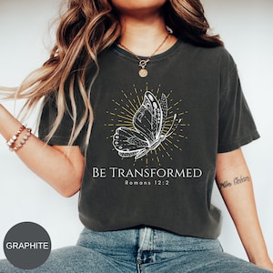 Può includere: Una maglietta grigia antracite con una grafica di farfalla bianca e il testo "BE TRANSFORMED Romani 12:2".