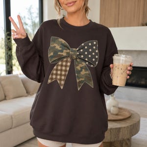 May include: A dark brown crewneck sweatshirt featuring a large central graphic of a bow. The bow is divided into four distinct sections, each with a different pattern: green and brown camouflage, white polka dots, brown gingham, and camouflage.