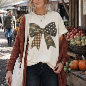 May include: A cream-colored short-sleeve t-shirt featuring a large graphic of a bow. The bow is divided into four sections with different patterns: camouflage, white polka dots on a dark background, brown gingham check, and solid olive green.