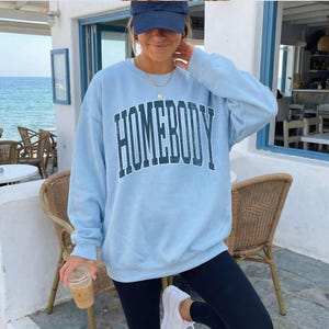 May include: A light blue crewneck sweatshirt featuring the word HOMEBODY printed in a dark blue, distressed, arched font across the chest. The garment has long sleeves, a ribbed collar, and ribbed cuffs, providing a relaxed and comfortable fit.