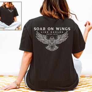 Könnte beinhalten: Schwarzes T-Shirt mit dem Text "SOAR ON WINGS LIKE EAGLES ISAIAH 40:31" und einer weißen Adlergrafik auf dem Rücken. Das Shirt ist dunkel und hat einen klassischen Rundhalsausschnitt. Das Model trägt das Shirt.