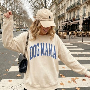 Może przedstawiać: Beżowa bluza z okrągłym dekoltem z napisem DOG MAMA w postarzanym niebieskim kroju czcionki, w zestawie z pasującą beżową czapką z daszkiem z małym ciemnym logo oraz czarną pikowaną torebką na ramię z łańcuszkiem.