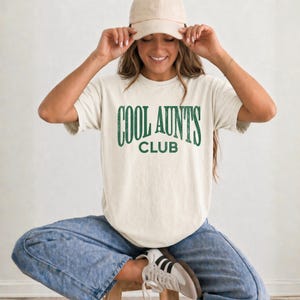May include: A cream-colored short-sleeve t-shirt featuring the dark green text COOL AUNTS CLUB printed in a distressed font across the chest. The shirt is paired with blue denim jeans, a matching cream baseball cap, and white sneakers.