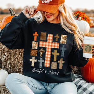 May include: A black long-sleeve sweatshirt featuring a large graphic print of multiple crosses in various patterns including orange gingham, plaid, leopard print, and floral. The text "Grateful & Blessed" is printed in white script below the crosses.