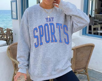 May include: A heather gray long-sleeve crewneck sweatshirt featuring the text YAY SPORTS in large, distressed blue block lettering across the chest. The garment has a relaxed fit with ribbed cuffs and a ribbed neckline, paired with black leggings.