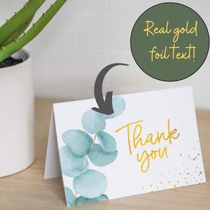 Może przedstawiać: Biała składana kartka z podziękowaniami ze złotym tekstem i wzorem liści eukaliptusa. Na kartce widnieje napis "Thank you" w złotym kolorze. Na zdjęciu widoczna jest również zielona roślina w białej doniczce.