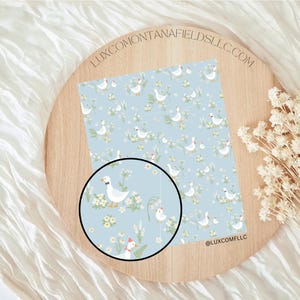 Puede incluir: Papel estampado azul claro con gansos blancos con pajaritas negras, rodeados de flores amarillas y follaje verde. El papel se muestra en una bandeja de madera con el texto "LUXCOMONTANAFIELDSLLC.COM" y "@LUXCOMFLLC".