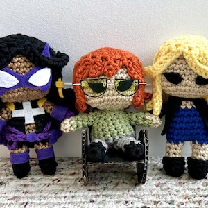 May include: Three crocheted dolls, one in a purple and black costume, one in a green shirt and grey pants, and one in a black jacket and blue dress. The dolls are all sitting on a brown surface.
