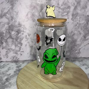 May include: A clear glass tumbler with a green and black Nightmare Before Christmas character design. The tumbler has a bamboo lid and a straw.